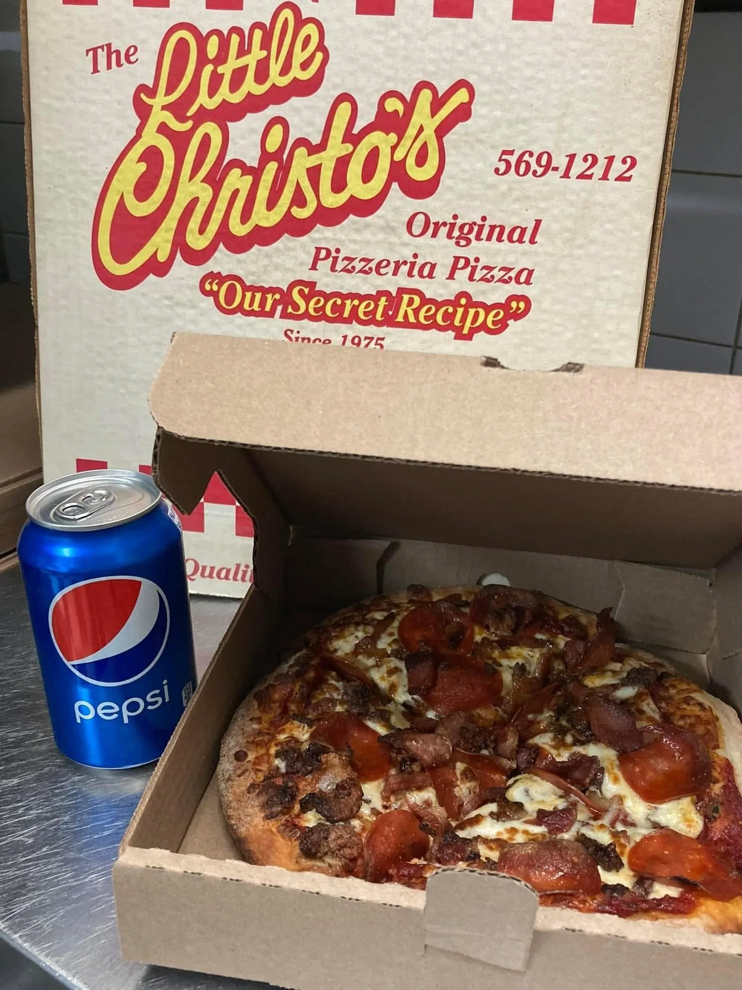 A Little Christo's pizza in the open printed box, the box lid showing the script logo, Our Secret Recipe and Since 1975, with a can of Pepsi beside it