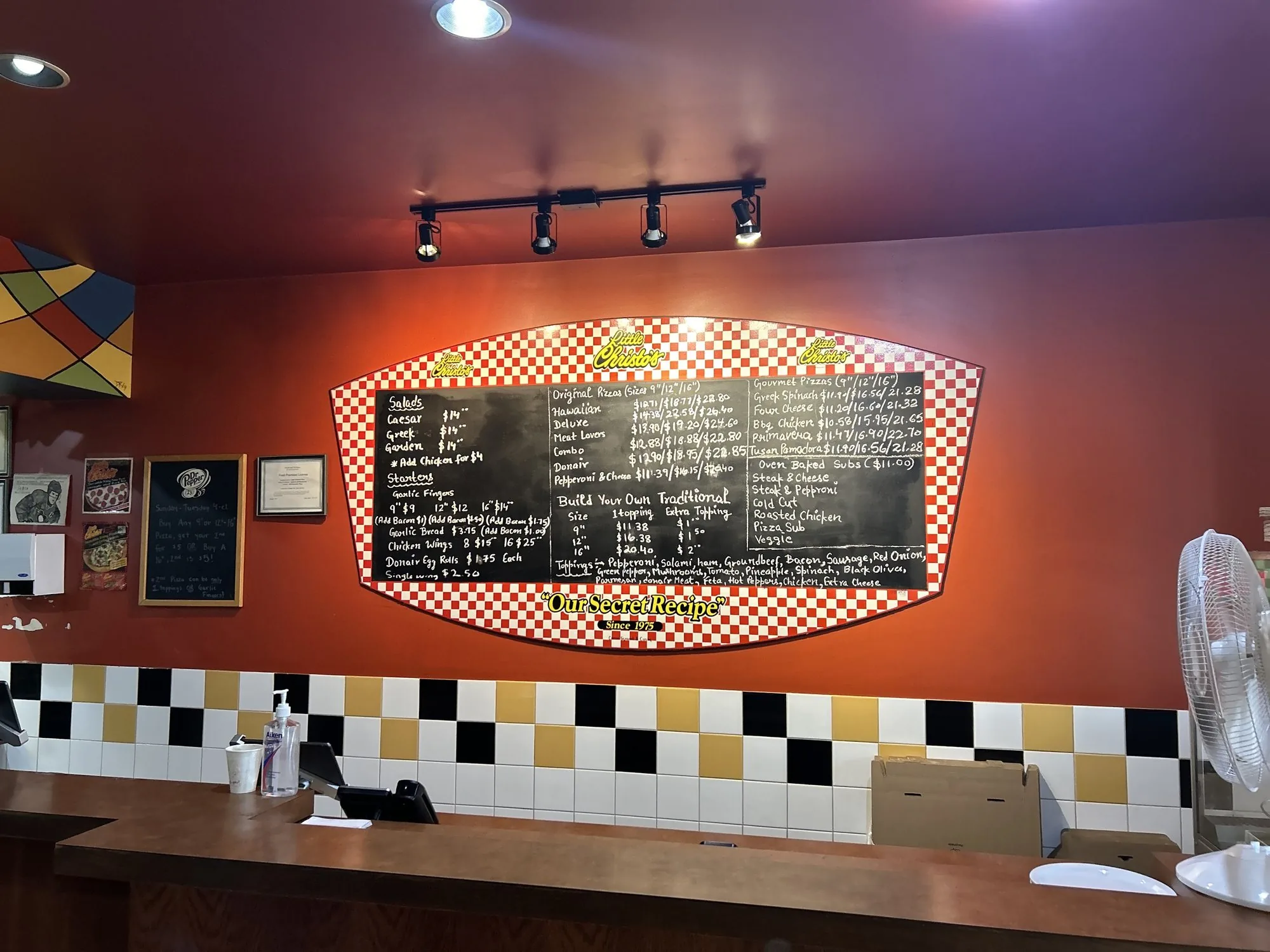 The red back wall of the shop with the oval checker-bordered menu board above the checker counter tile