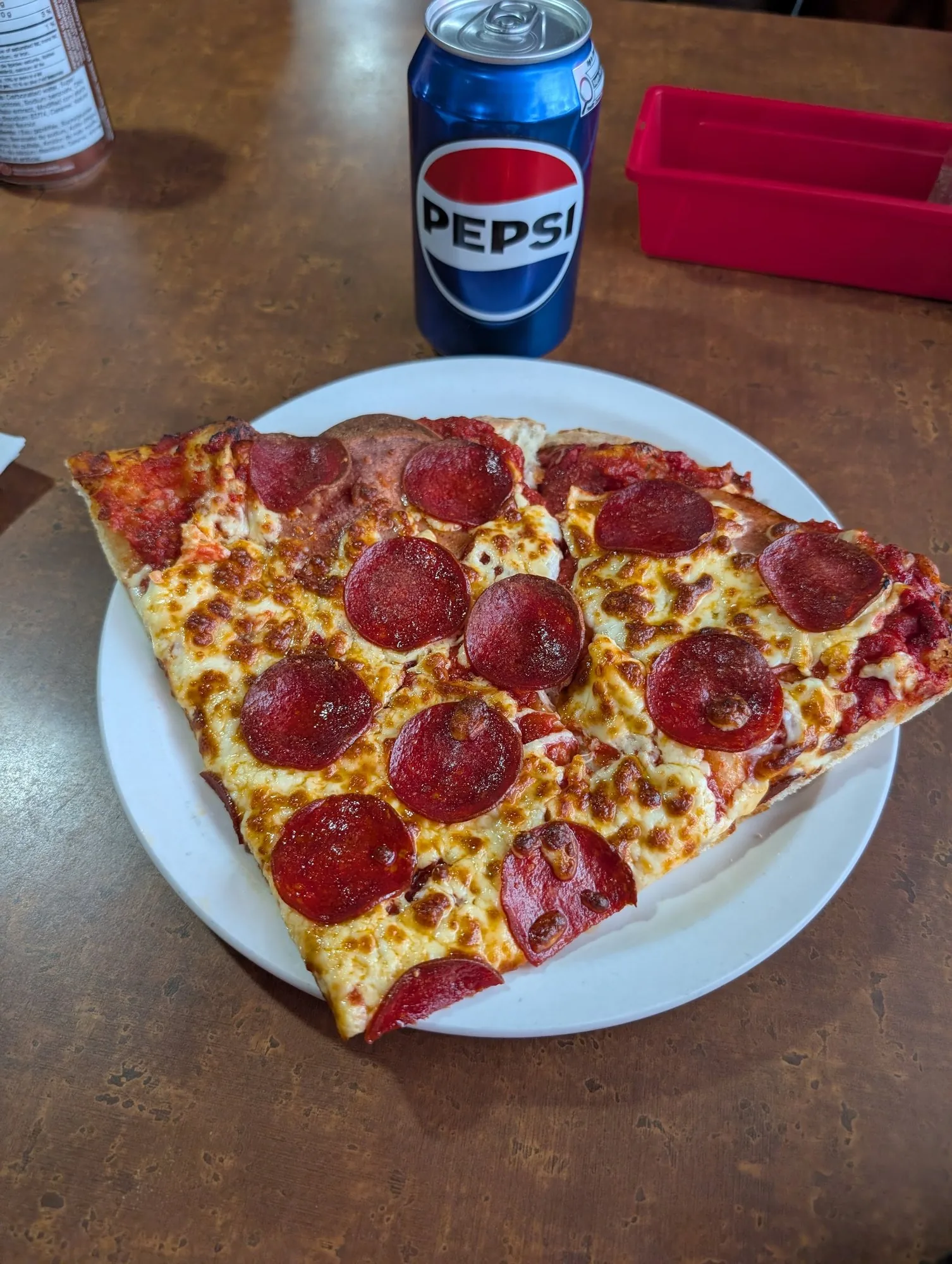 A big pepperoni slice on a white plate with a Pepsi on the dining table